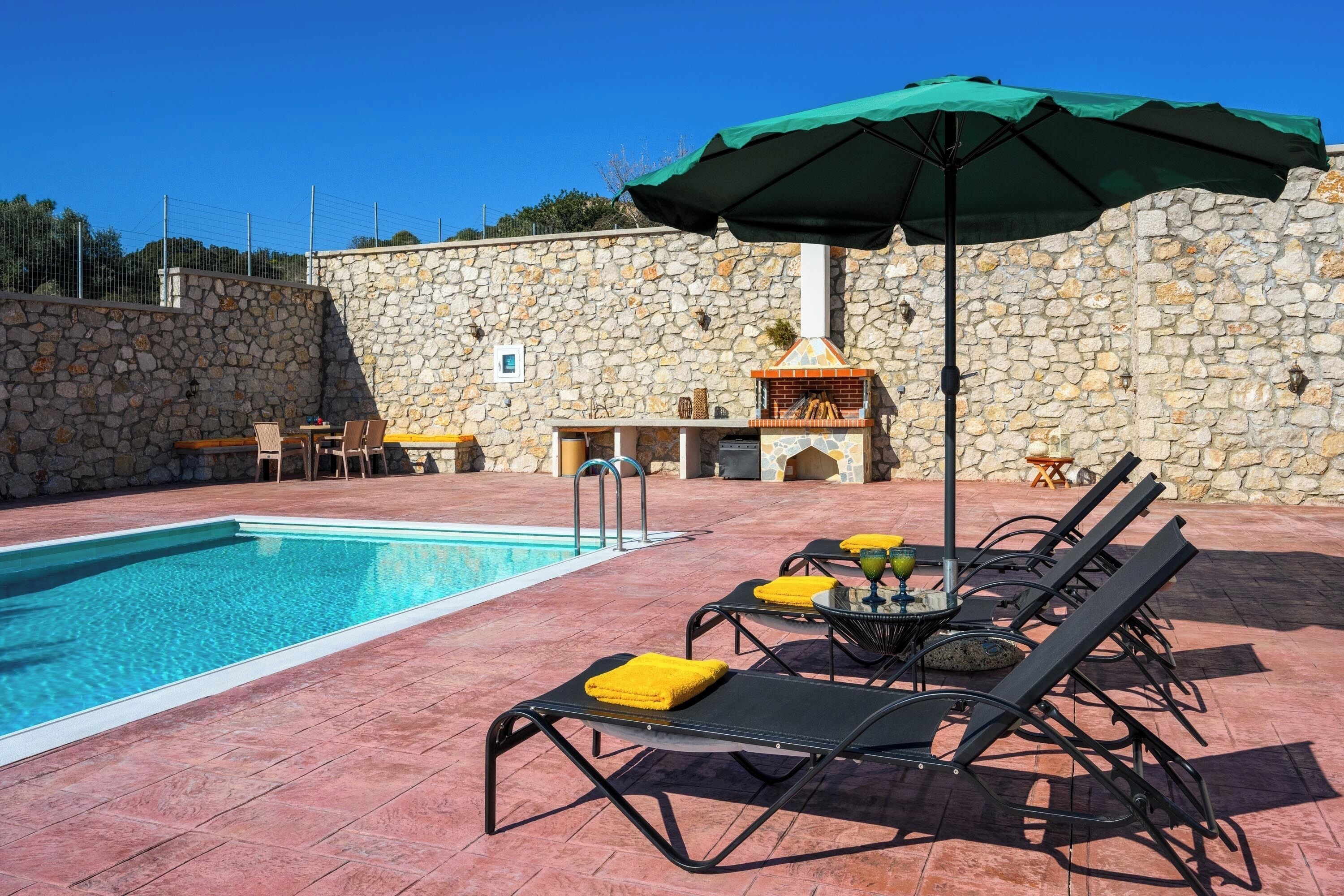 Une piscine avec des chaises longues et une table, un mur en pierre et un espace barbecue.
