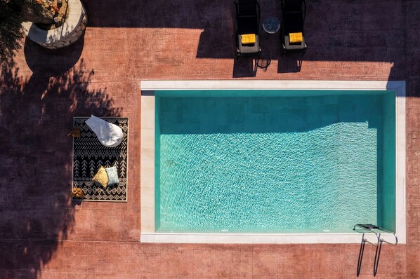 An aerial view of a rectangular swimming pool with a clear blue surface, surrounded by a tiled deck with a patterned mat and a small potted plant.