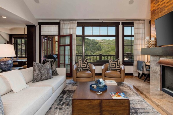 A modern living room with a sofa, two armchairs, a coffee table, and a fireplace. Large windows offer a view of a mountainous landscape.