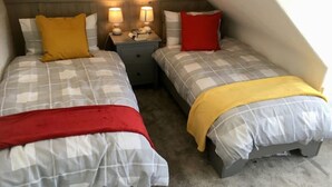 A small, cozy bedroom with two beds, each with a distinct color accent pillow, a nightstand with lamps, and a simple headboard.