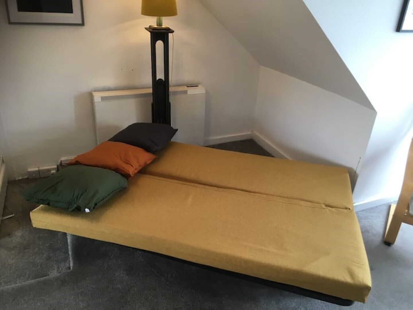 A small, single-bed room with a plain mattress, a standing lamp, and a radiator.
