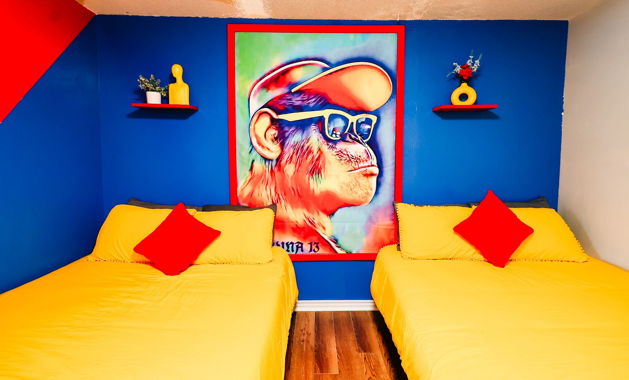 A room with two yellow beds, red pillows, a large colorful poster on the wall, and small shelves with decorative items.