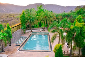 A rectangular pool with a stone deck, surrounded by palm trees and greenery, with mountains in the background. - The Pure Nature Hotel (Rajgarh)