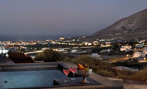 A table with two glasses of pink drink and a fruit bowl, overlooking a coastal town at dusk. - ALTURA RESIDENCES BY K&K (Santorini)