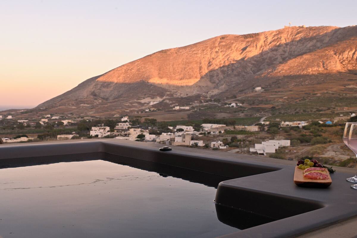 A poolside view with a mountain backdrop, a table with a glass of wine and food, and a chair.