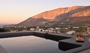 A poolside view with a mountain backdrop, a table with a glass of wine and food, and a chair. - ALTURA RESIDENCES BY K&K (Santorini)