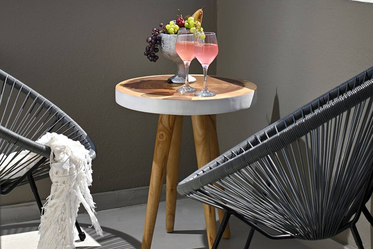 A terrace with a table set for two, featuring a fruit bowl and two glasses of pink beverage. The terrace overlooks a coastal landscape with buildings and a clear sky.