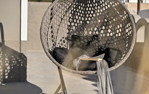 A wicker chair with a cushion and a table, positioned on a balcony with a concrete floor and a white wall in the background. - ALTURA RESIDENCES BY K&K (Santorini)