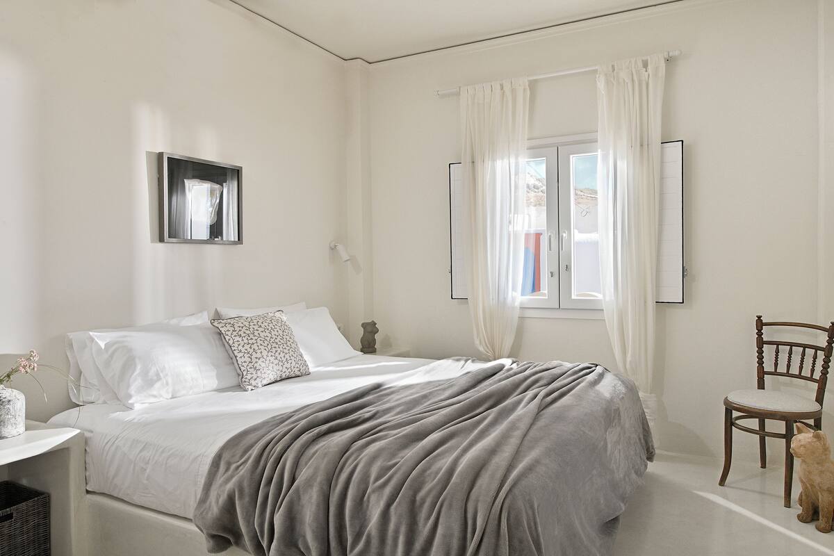 A bedroom with a bed, a chair, a window with curtains, and a flat-screen TV mounted on the wall.