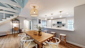 A modern kitchen with white cabinets, a wooden dining table, and white chairs. There is a staircase with white railings leading to another level.