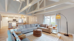 A spacious living room with a large L-shaped sofa, a round wicker coffee table, and a floor lamp. A kitchen with white cabinets and a dining area with a table and chairs are visible in the background.