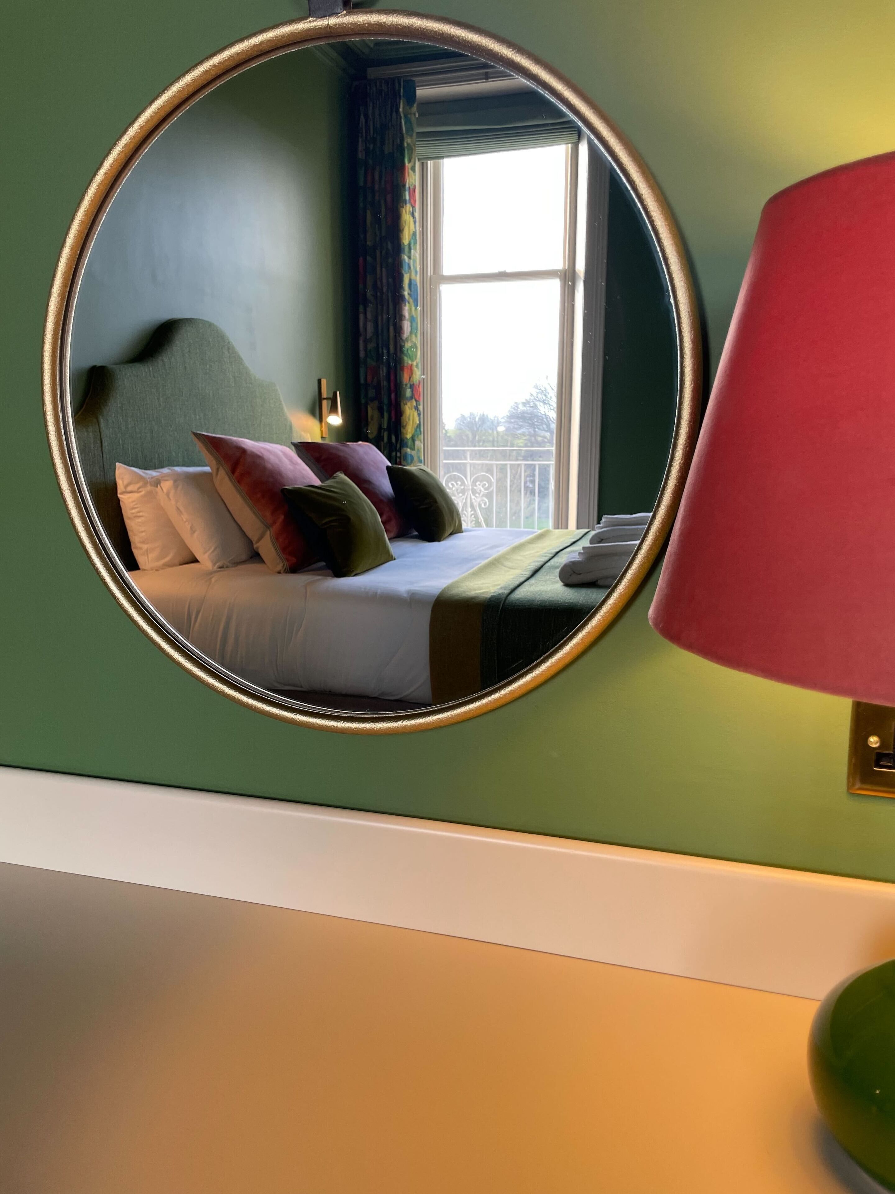 Un dormitorio con un espejo redondo, una cama con almohadas, una mesita de noche y una ventana con cortinas.