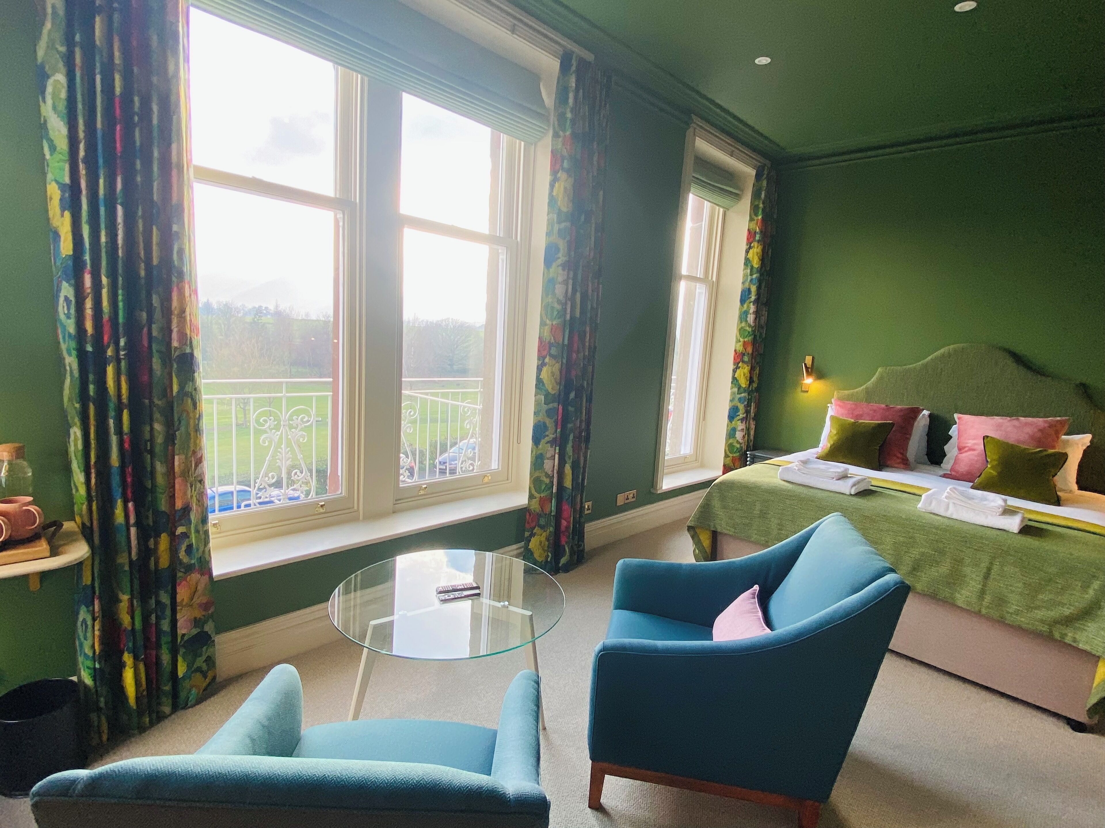 A room with a bed, two chairs, a glass table, and large windows with curtains.