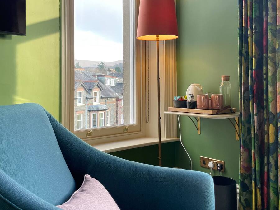 A room with a blue armchair, a red floor lamp, a window with a view of buildings, and a shelf with items.