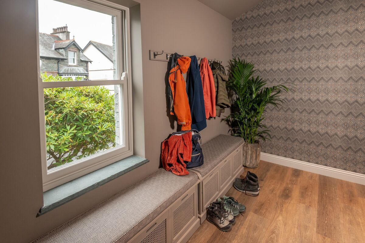 Une pièce avec un banc près d’une fenêtre, un porte-manteau mural avec des vestes, une plante en pot et un mur décoré d’un motif.