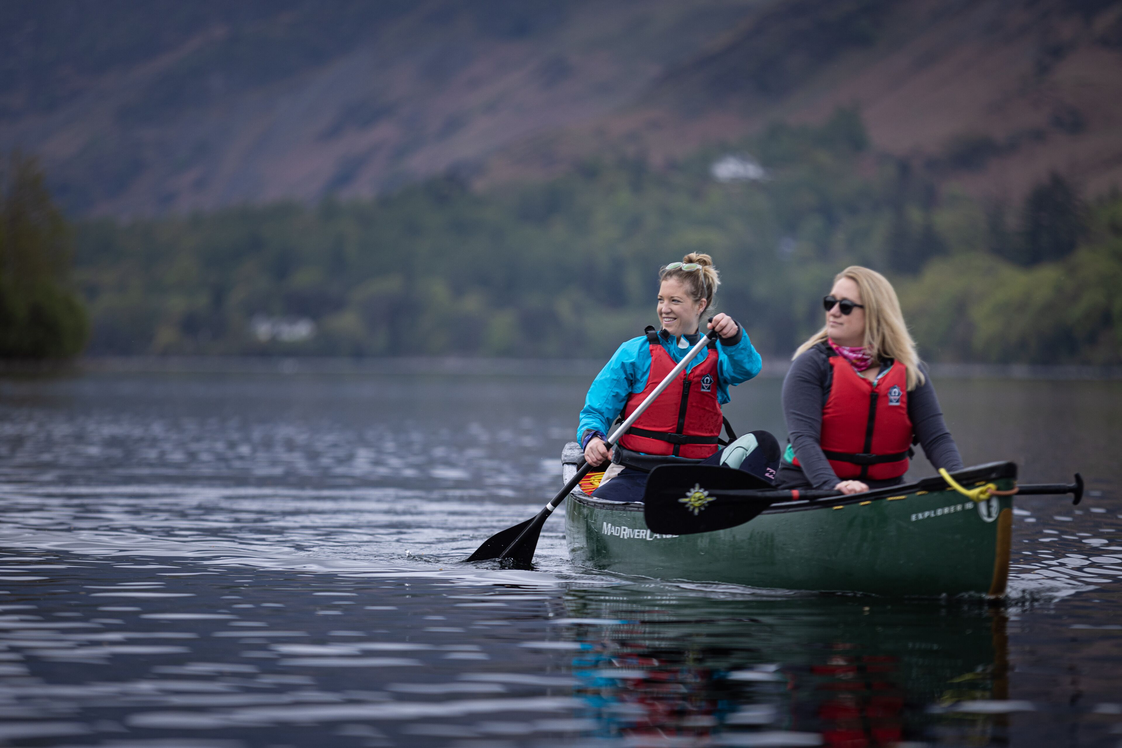 Two people in a green canoe on a calm lake with mountains in the background.