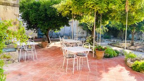 Une terrasse extérieure avec des tables et des chaises en métal blanc, entourée de plantes en pot et d’arbres.