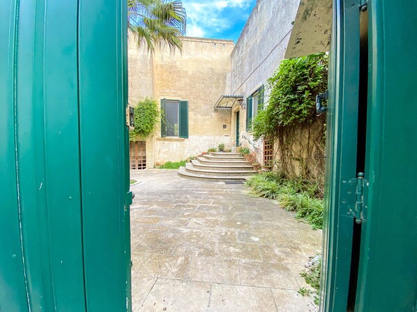 Une cour avec des marches en pierre menant à l’entrée d’un bâtiment, entourée de verdure et sous un ciel dégagé.