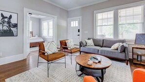 A spacious living room with a gray sofa, a coffee table, and a brown leather chair. There is a ceiling fan, large windows with blinds, and a framed picture on the wall. - Adorable Home. Close to Everything. Great Location (Charlotte)