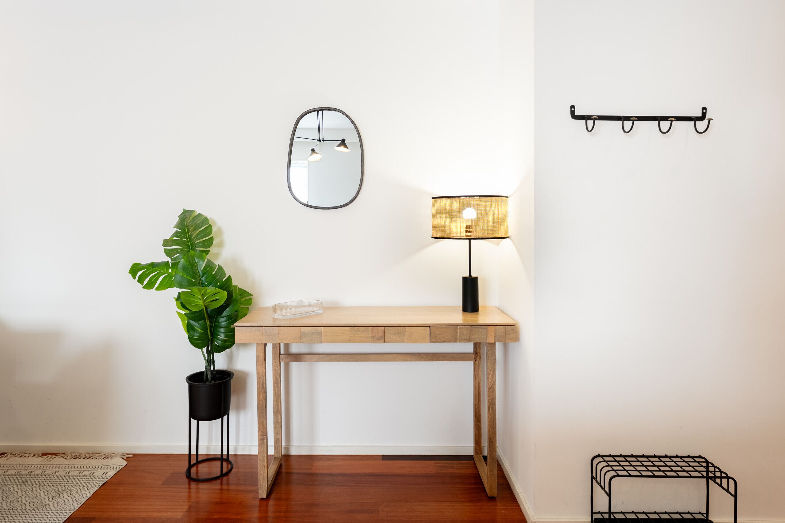 A wooden desk with a lamp, a potted plant, a mirror, and a metal rack on a white wall.