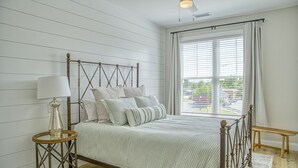 Un dormitorio con estructura de cama de madera, una mesita redonda con lámpara, un ventanal grande con persianas y un ventilador de techo.