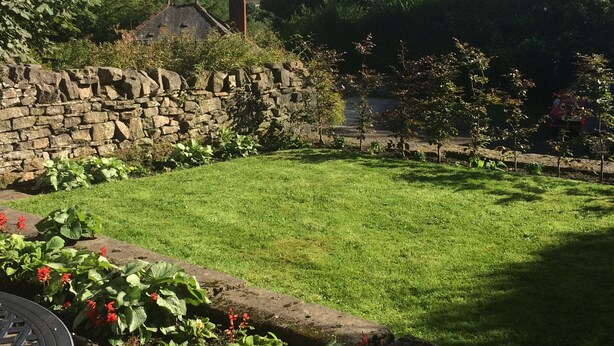 A stone wall, a grassy yard, and a building with a chimney in the background. - Hawkyard's Cottage (Oldham)