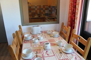 A dining area with a table set for four, surrounded by wooden chairs. A window with a view of a kitchen is above the table.
