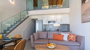 A modern living room with a sofa, dining table, and chairs. A staircase leads to an upper level. - OceanEye Suites - Fortuna (Panormo)