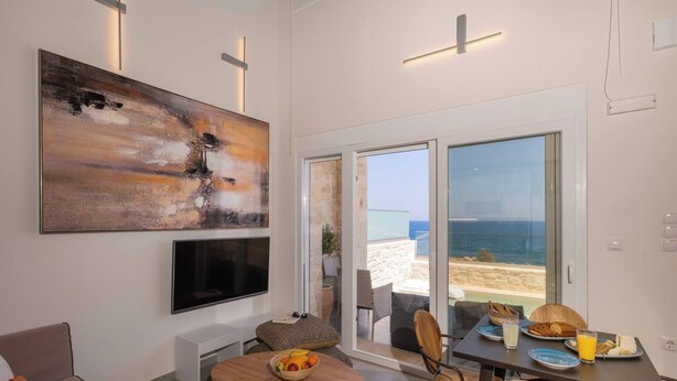 A modern living room with a large abstract painting, a flat-screen TV, and a dining area with breakfast items. - OceanEye Suites - Fortuna (Panormo)