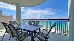 A balcony with a table and chairs, overlooking a beach with clear blue water and a pier.