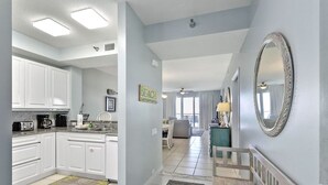 A modern kitchen with white cabinets, a dining area with a wooden bench, and a hallway leading to a living room.
