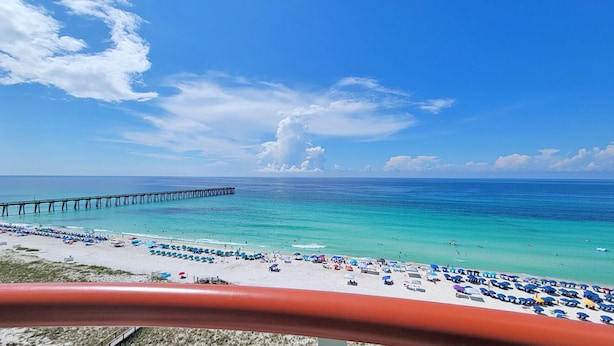 A beach with a pier extending into the ocean, clear turquoise water, and numerous beach umbrellas.