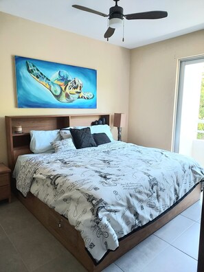 A bedroom with a bed, a ceiling fan, a painting of a turtle, and a window. - Stylish Jungle Condo, Walking Distance to Beach (Akumal)