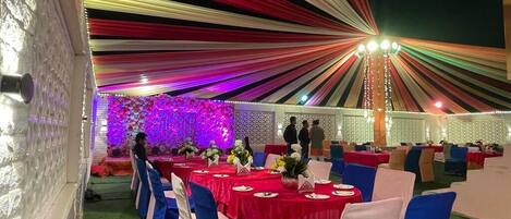 A banquet hall with round tables covered in red tablecloths, blue chairs, and a decorative backdrop with purple lighting.