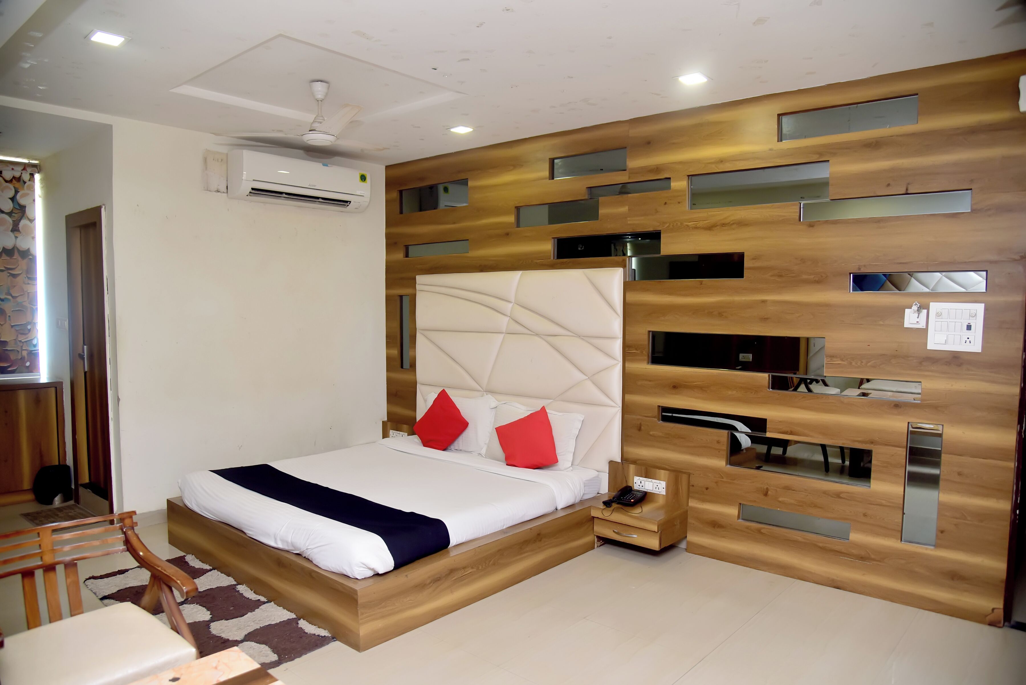 A modern bedroom with a wooden headboard, a bed with white bedding and red pillows, a bedside table with a phone, and a ceiling fan.