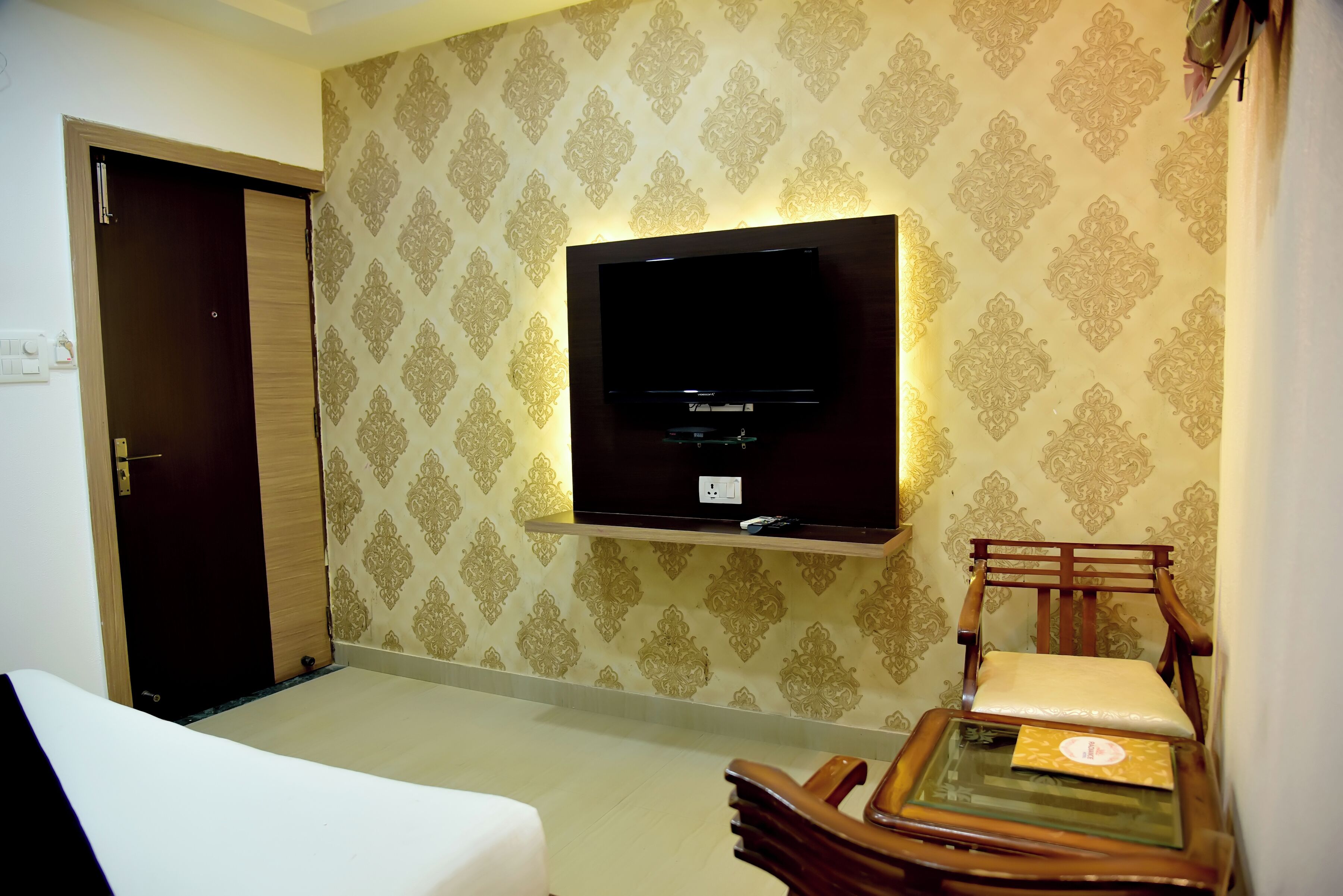 A hotel room with a flat-screen TV mounted on the wall, a wooden chair with a cushion, and a bed with a white cover.