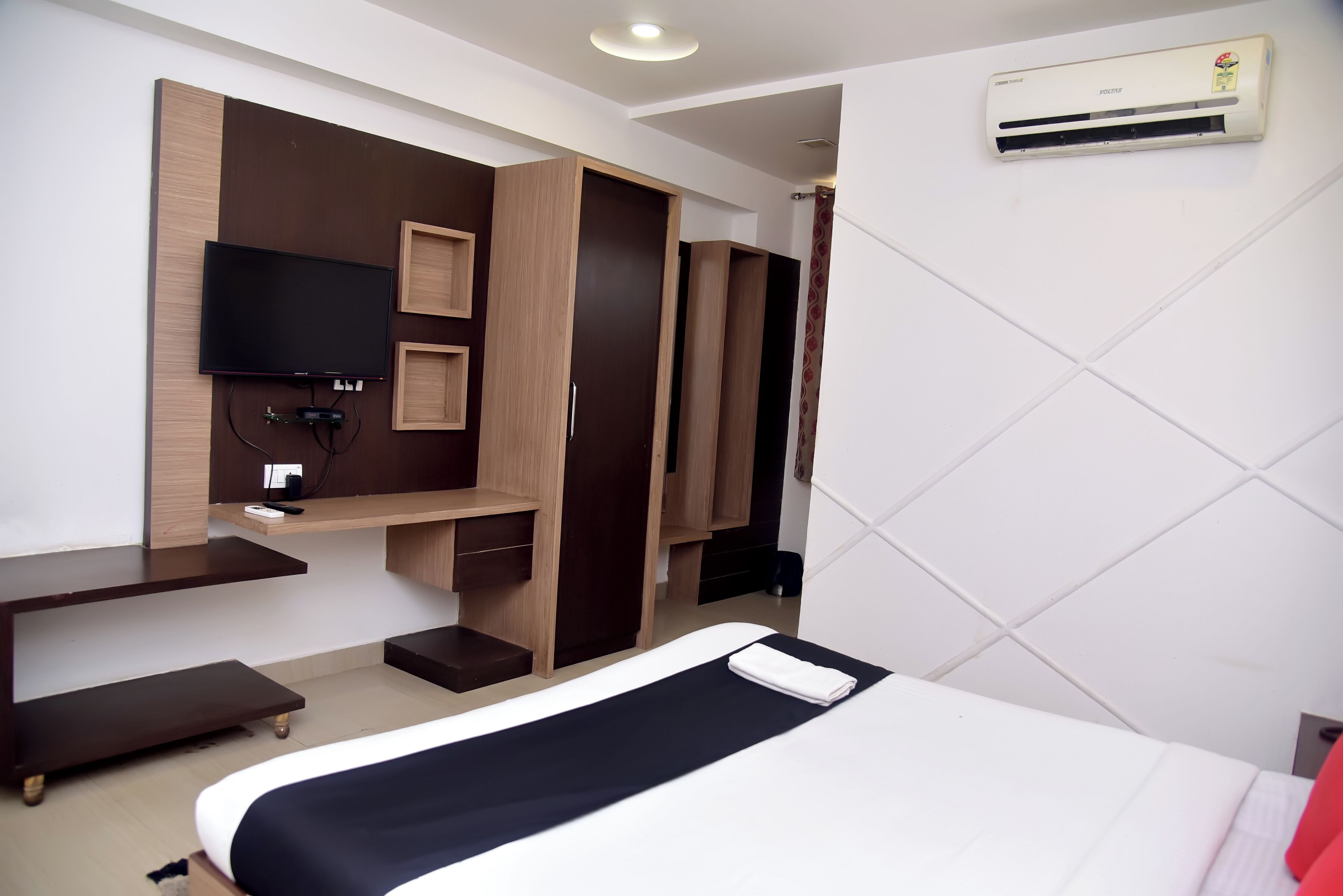A modern hotel room with a bed, a flat-screen TV mounted on a wooden panel, a desk with a chair, and built-in wardrobes.