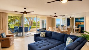 A modern living room with a large blue sofa, a brown armchair, and a dining area with wooden furniture. - Exceptional Oceanview at Royal Mauian | Rare 3 Bedroom and Oceanfront Living (Kihei)