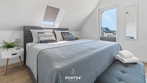 Un dormitorio moderno con un cabecero tapizado en gris, una colcha estampada en gris, una mesita de noche blanca con una planta, y un ventanal con vistas a edificios.