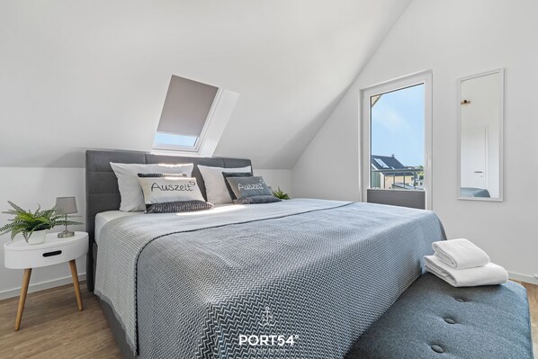 A modern bedroom with a grey upholstered headboard, a grey patterned bedspread, a white bedside table with a plant, and a large window with a view of buildings.