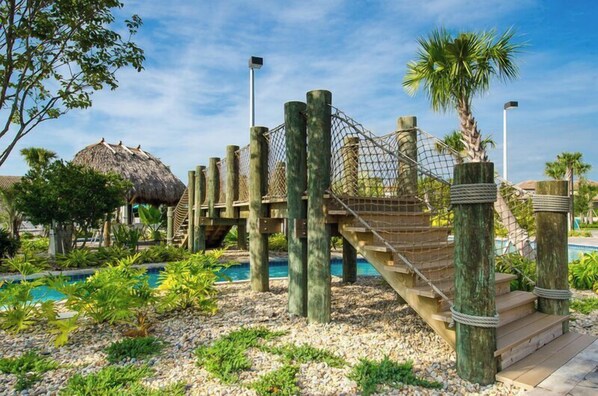 Un puente colgante sobre una zona de piscina con una estructura de techo de paja al fondo.