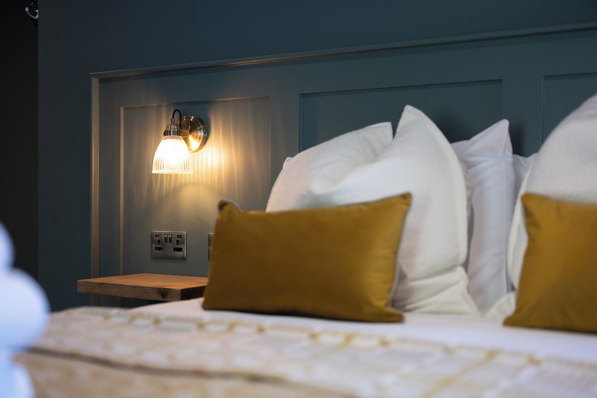 Una habitación de hotel con cama, almohadas, mesita de noche y una lámpara en la pared.