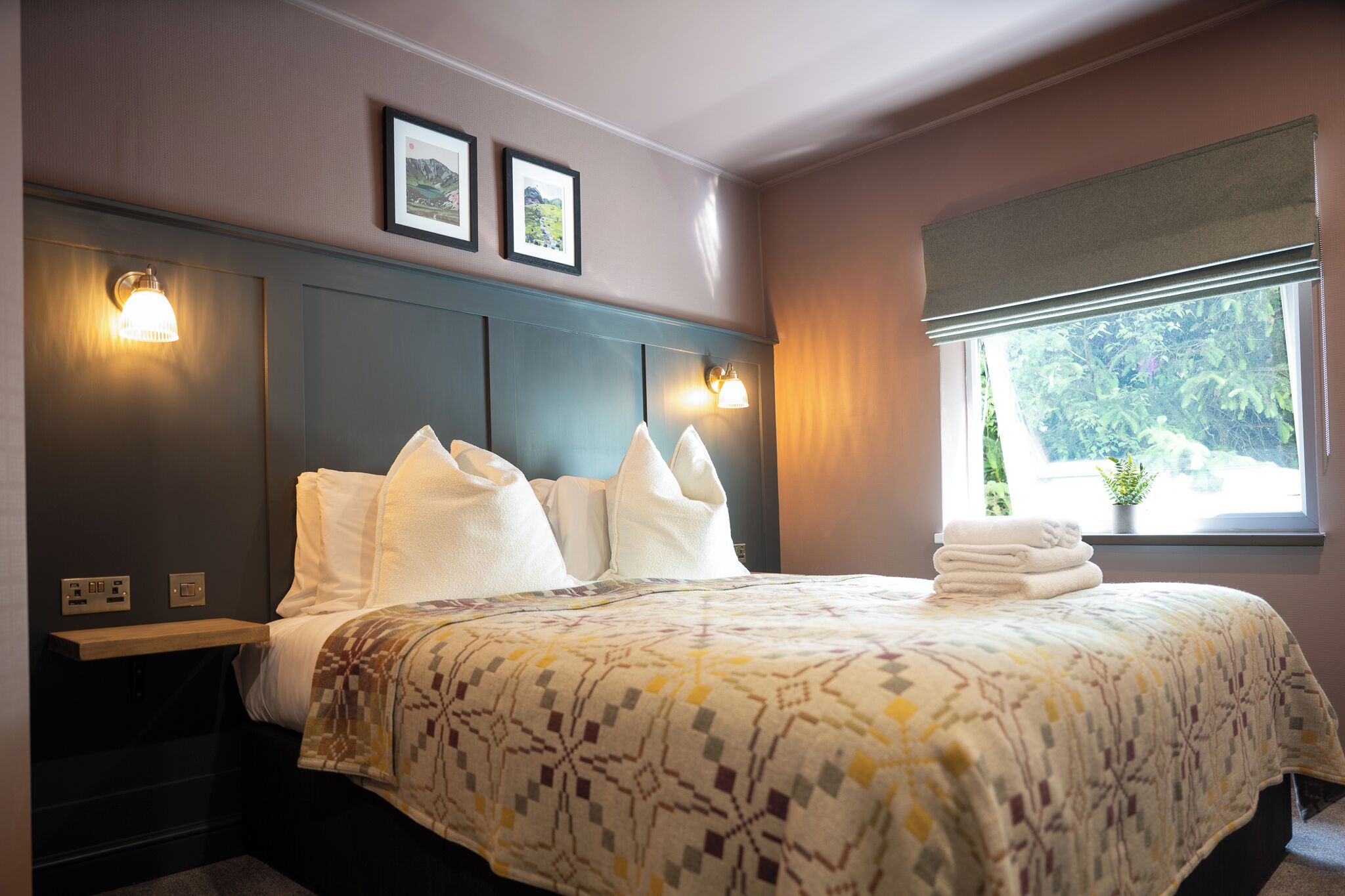 A neatly made bed with a patterned bedspread, two wall-mounted light fixtures, a window with a view of greenery, and a framed picture on the wall.