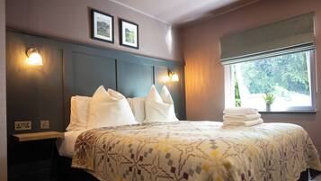 A neatly made bed with a patterned bedspread, two wall-mounted light fixtures, a window with a view of greenery, and a framed picture on the wall.