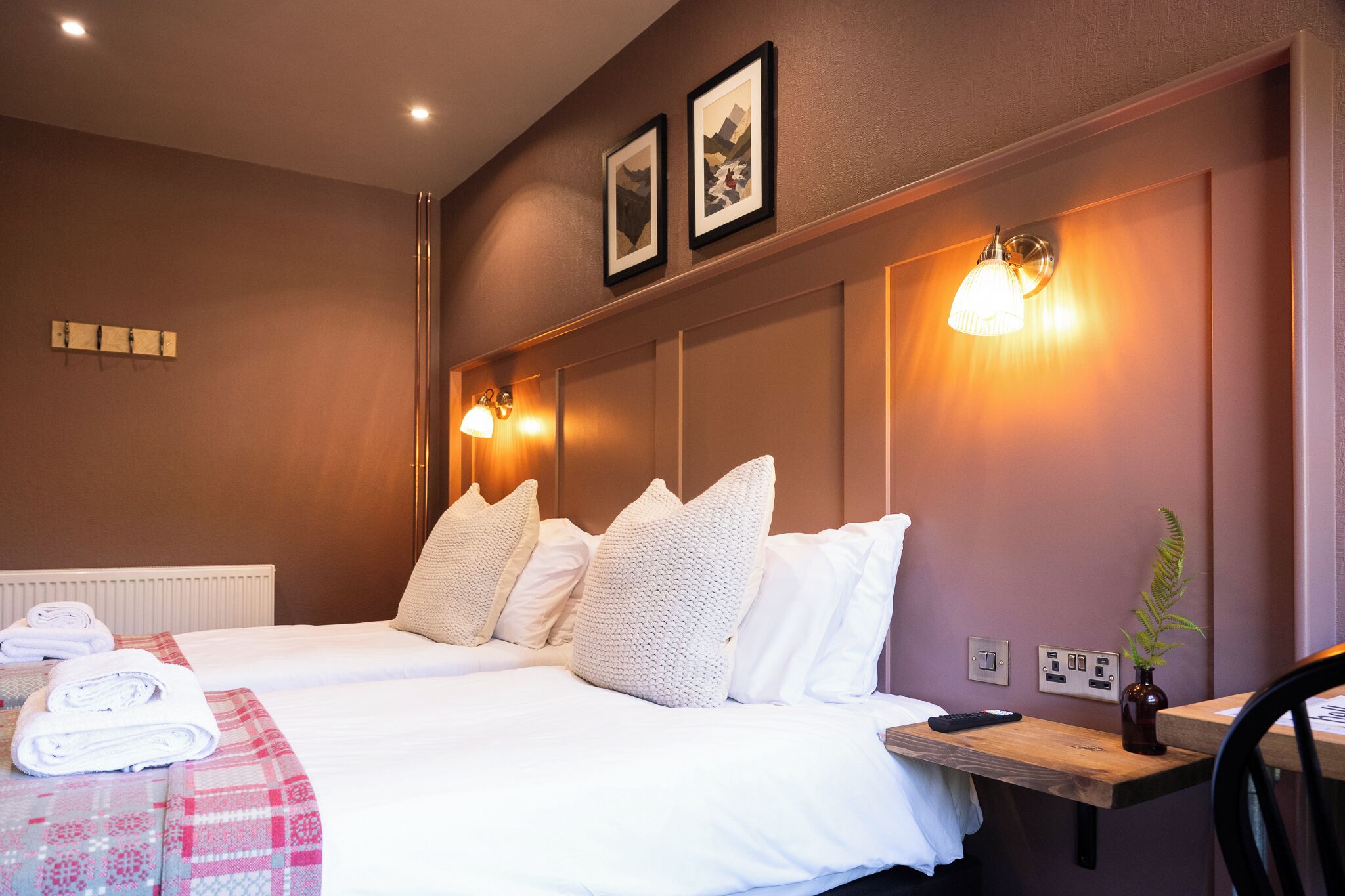 Une chambre d’hôtel avec deux lits, une table de chevet en bois, une chaise, un radiateur, des appliques murales et une petite plante.