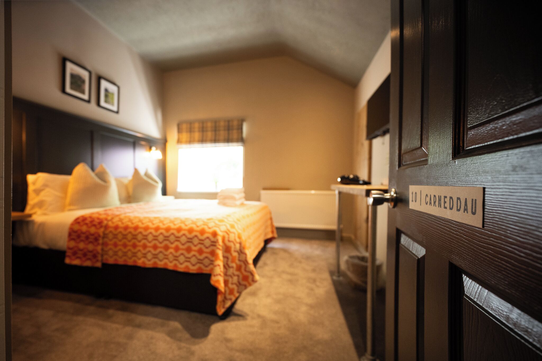 Une chambre d’hôtel avec un lit, une fenêtre ornée d’un rideau à carreaux, une porte avec une plaque nominative et un radiateur.