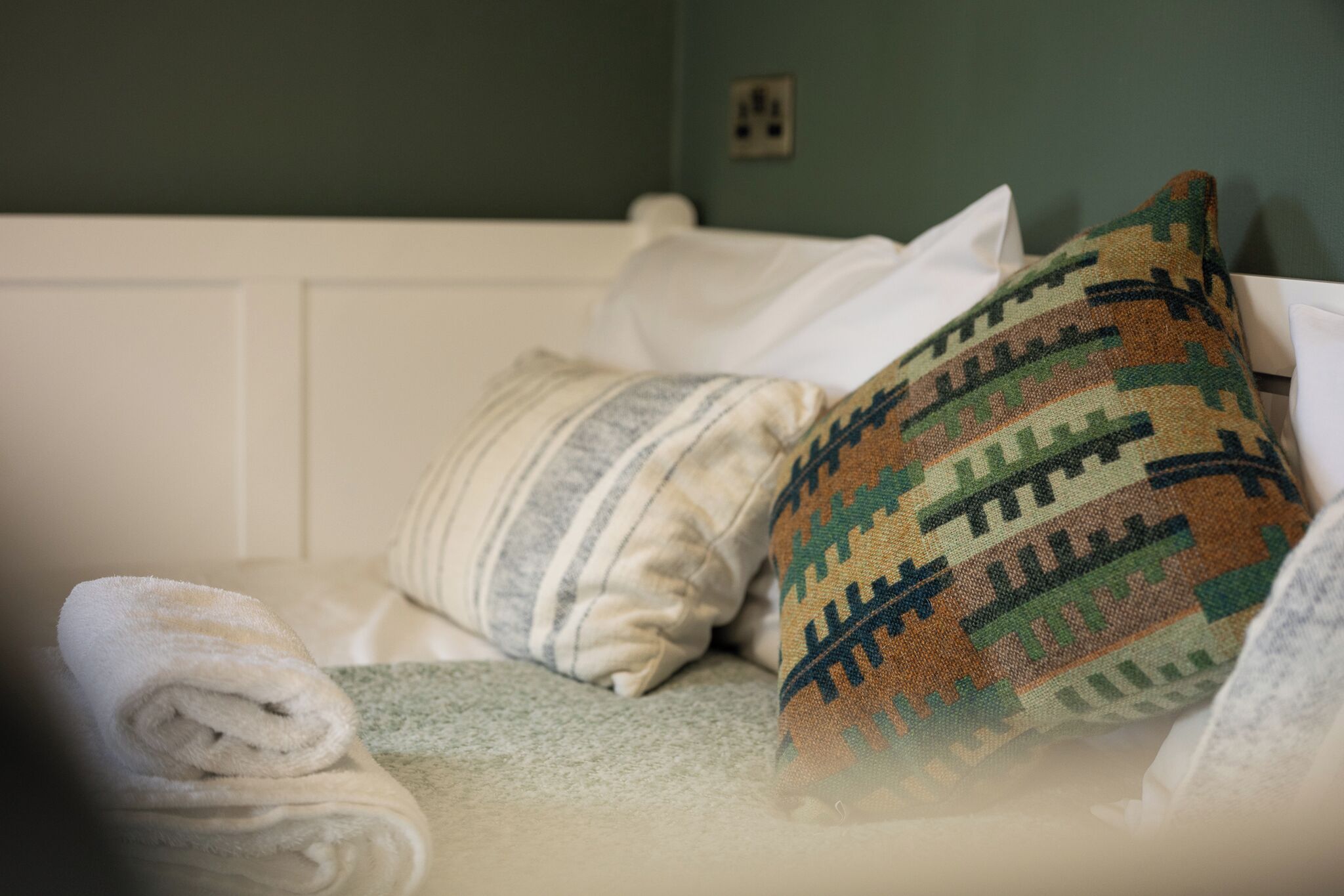 Una cama con sábanas blancas, una toalla enrollada y una almohada estampada.