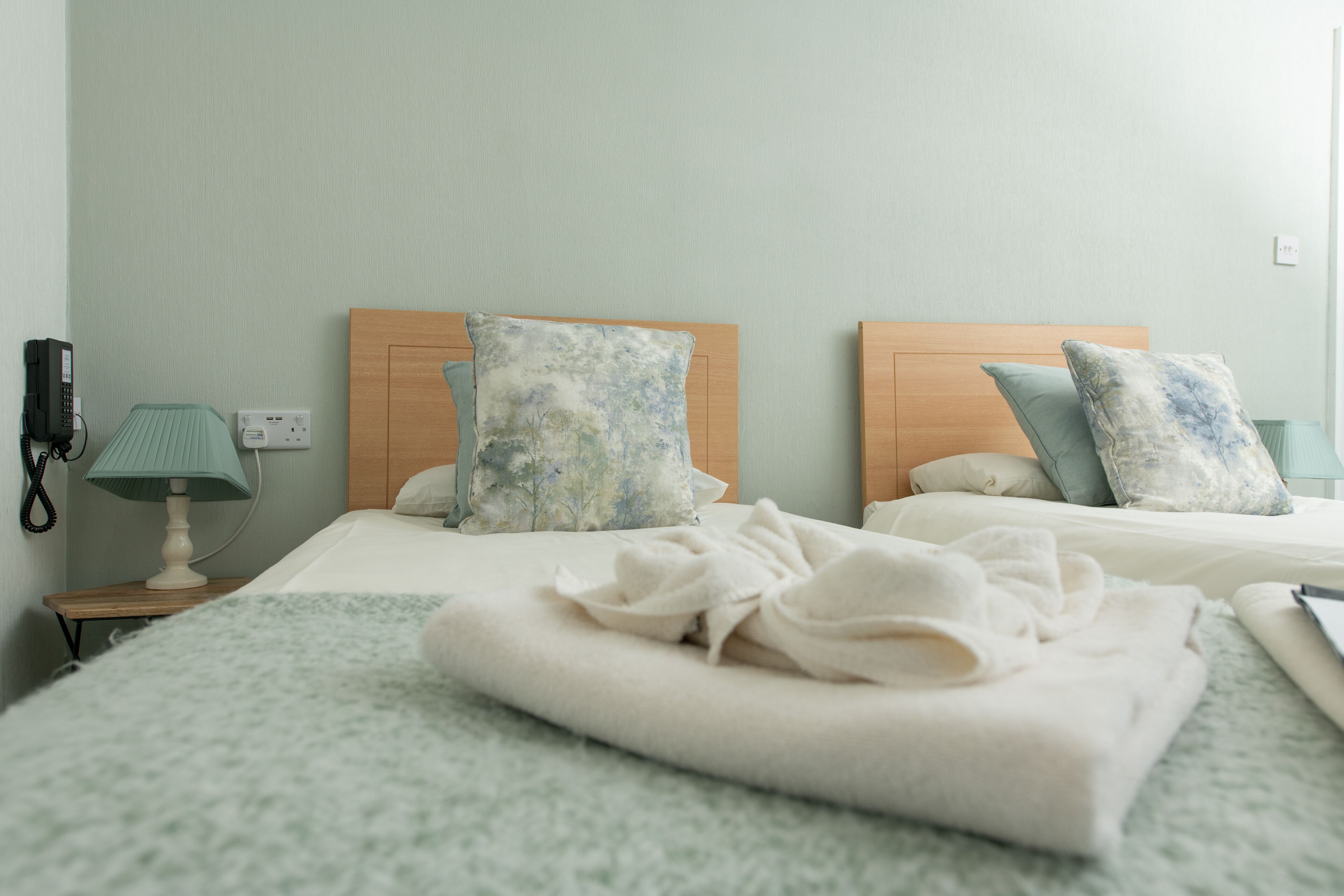 A hotel room with two beds, each with a wooden headboard and floral-patterned pillows. There is a bedside table with a lamp and a phone on the left side.