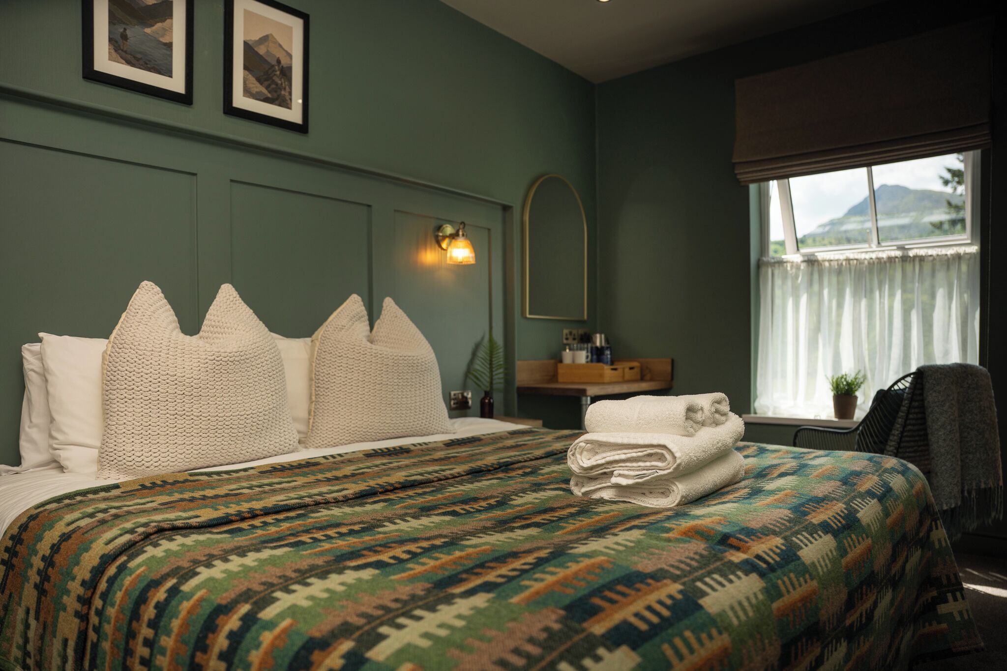 A neatly made bed with a patterned bedspread, a bedside table with a lamp, a window with curtains, and framed pictures on the wall.
