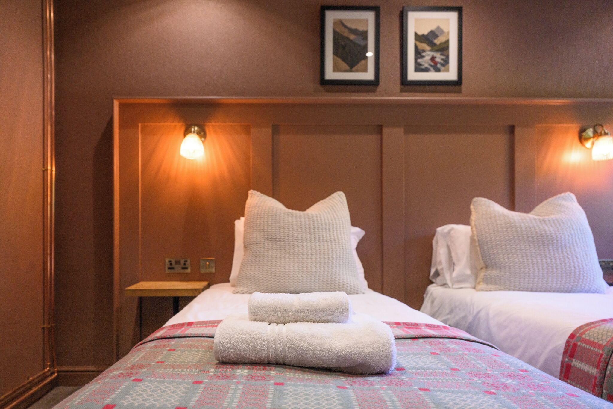 A hotel room with two beds, a nightstand, and wall-mounted lamps.